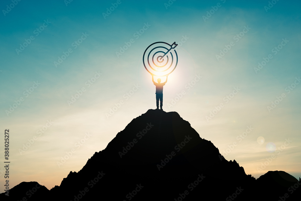 Silhouette of businessman holding target board on the top of mountain with over blue sky and sunlight. It is symbol of leadership successful achievement with goal and objective target.