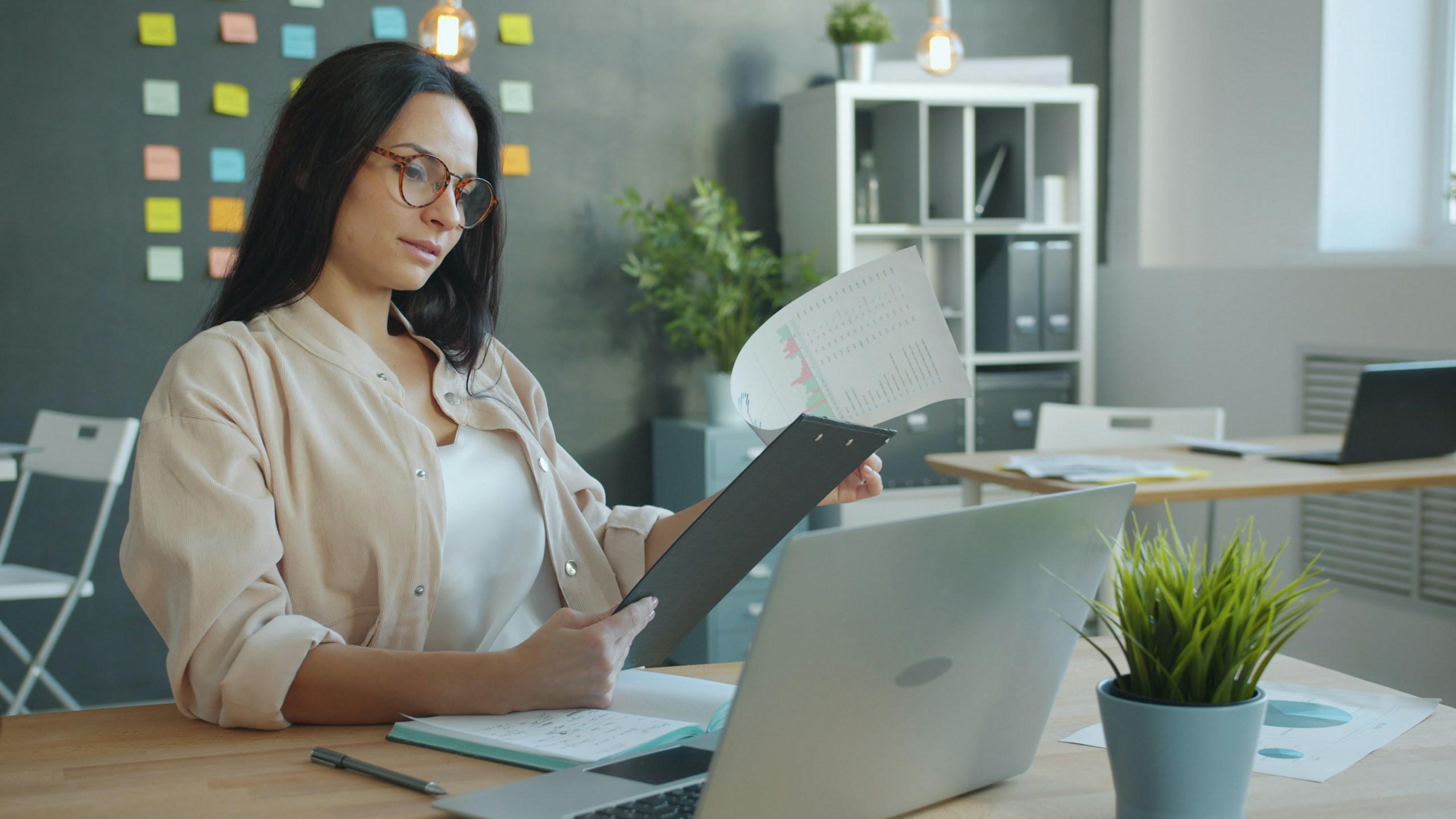Businesswoman attractive young woman in elegant clothing is looking at document and laptop screen focused on work in office. Business and information concept.